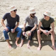 Three men sitting at the water’s edge wearing matching cream bucket hats with orange oh my hats! embroidery.