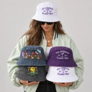 Woman wearing a white embroidered bucket hat and holding four hats in washed blue, purple, charcoal gray, and white.