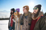Four friends wearing black, purple, and brown embroidered beanies in a snowy mountain setting.