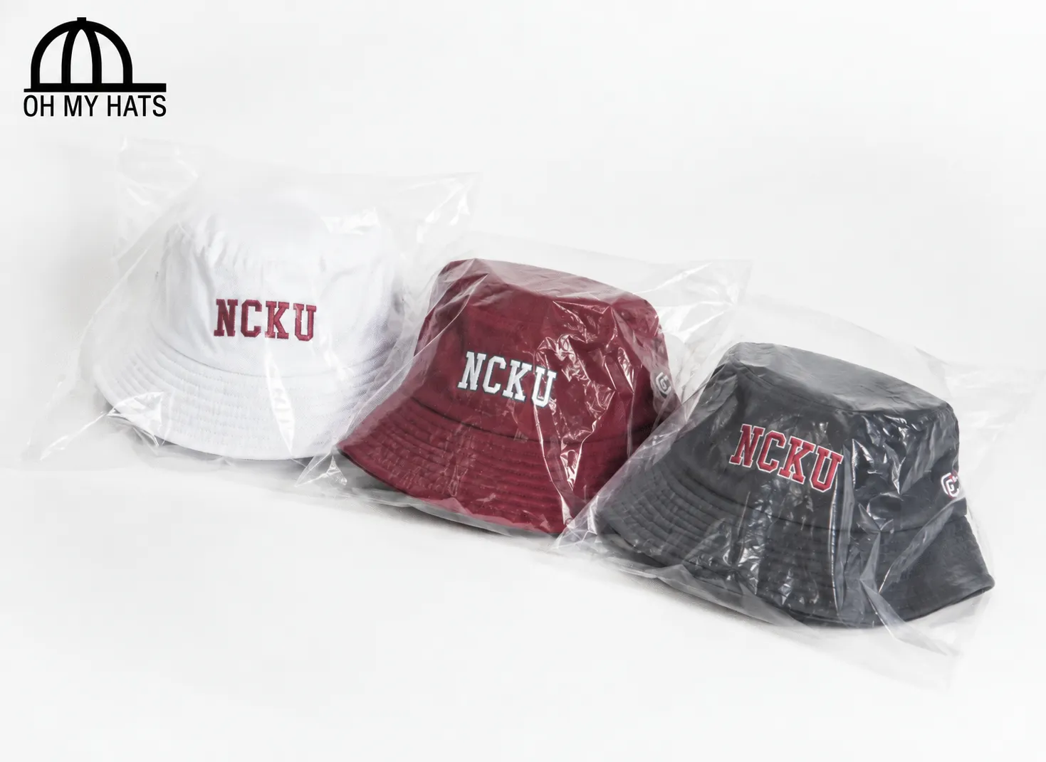 Three individually packaged bucket hats in white, burgundy, and charcoal with NCKU embroidery.
