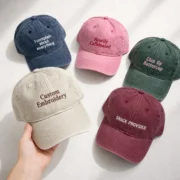 Five washed dad hats in navy, pink, dark green, beige, and burgundy with different embroidered phrases.