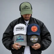 Man wearing an olive green OH MY HATS! embroidered trucker hat and holding four embroidered caps against a gray background.