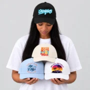 Model wearing a black OHMYHATS five-panel printed baseball hat and holding three colorful printed hats against a neutral gray background.