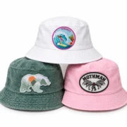 Three patch bucket hats arranged in a triangular stack: white with a Hawaii surfing patch, green with a mountain bear patch, and pink with a Mothman patch.