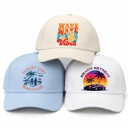 Three solid-color five-panel baseball hats in cream, light blue, and white, stacked with colorful beach-themed printed designs.