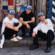 Three chefs wearing blue and white foam trucker hats with KITCHEN CREW patches, seated outside a graffiti-covered doorway.