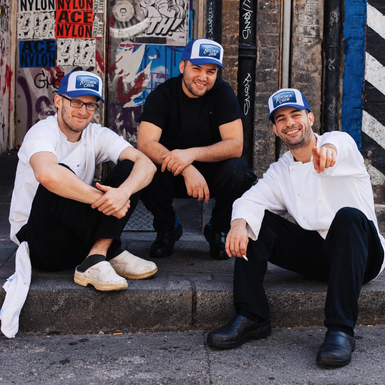 Three chefs wearing blue and white foam trucker hats with KITCHEN CREW patches, seated outside a graffiti-covered doorway.