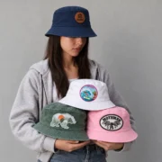 Model wearing a navy bucket hat with a brown logo patch and holding white Hawaii, green bear, and pink Mothman patch bucket hats.
