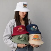 Model wearing a white palm-print bucket hat and holding four custom printed bucket hats in burgundy, navy, sage green, and beige against a light gray wall.