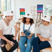 Three people wearing white palm-print bucket hats while reviewing a tablet together in a bright art studio.
