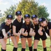 Five women in black sportswear wearing matching black TEAM SPIRIT patch baseball hats and giving thumbs-up outdoors.
