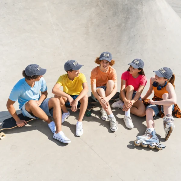 Five friends wearing washed slate blue dad hats with custom SKATE CREW dice patches while relaxing at a sunny skatepark.