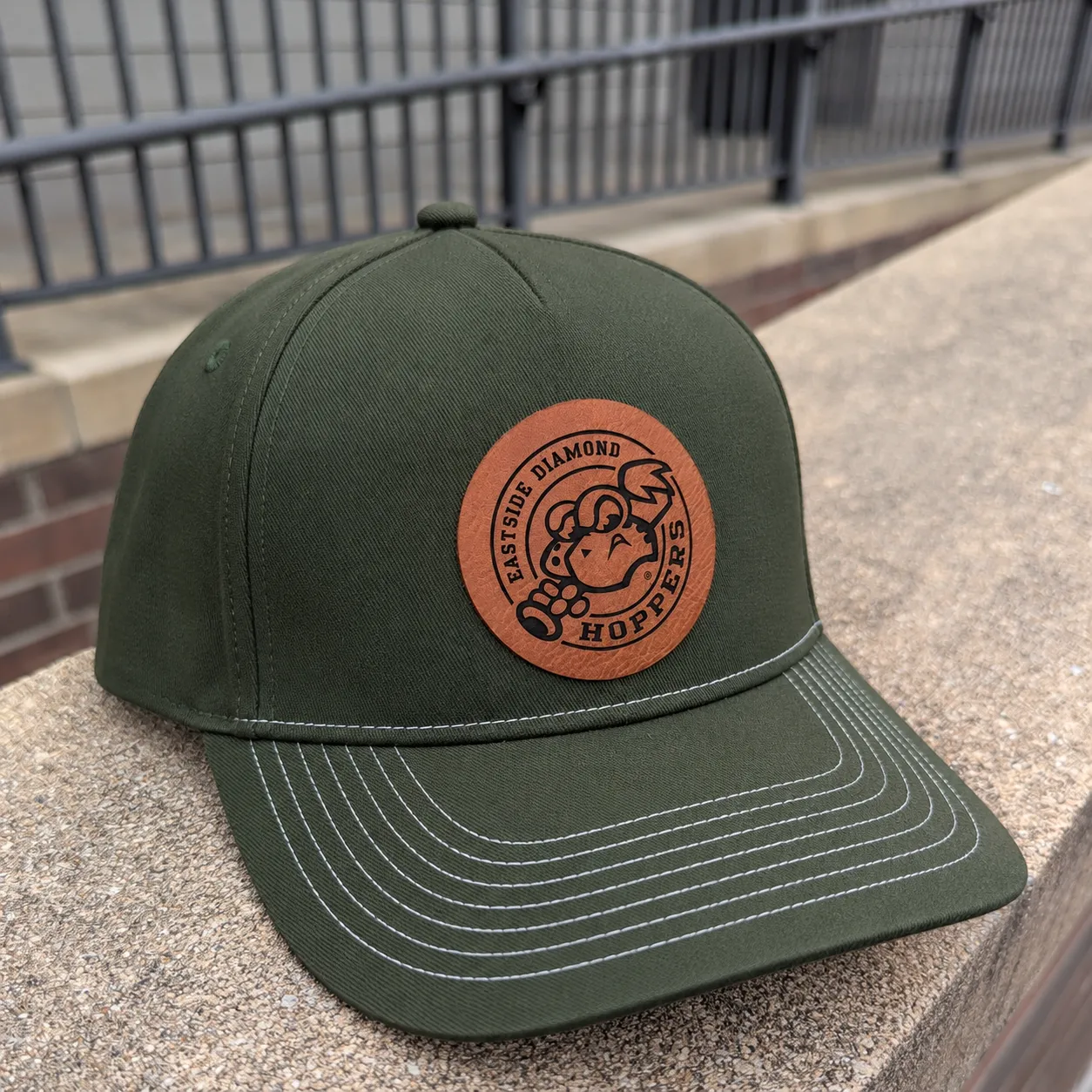 Olive green five-panel baseball hat with a brown Eastside Diamond Hoppers patch resting on a stone ledge outdoors.
