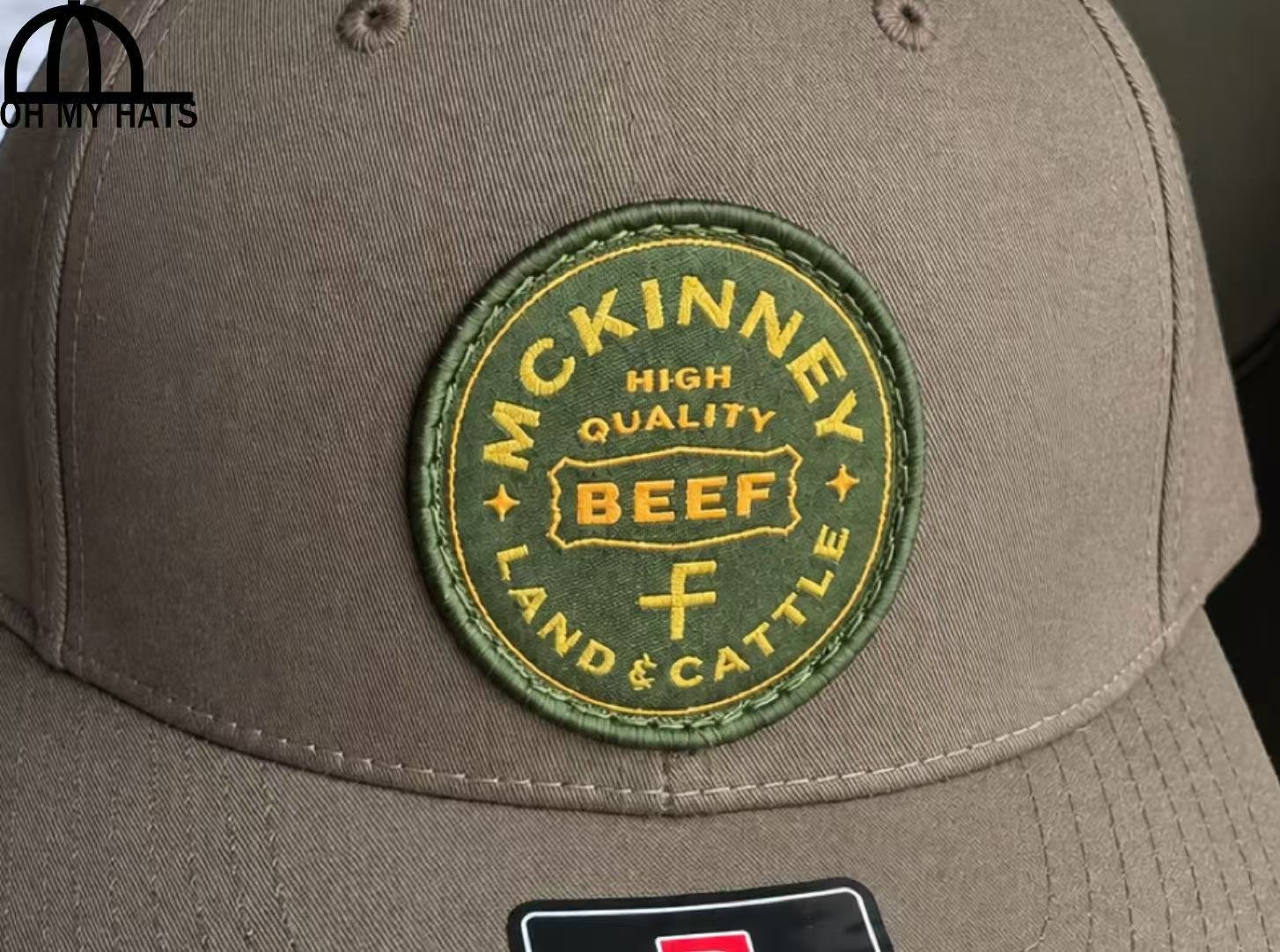 Hand holding a taupe five-panel baseball hat with a green and yellow McKinney Land & Cattle patch indoors.
